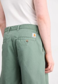 Pantalones de algodón verdes con corte recto, que cuentan con bolsillos traseros, un botón marrón y una etiqueta blanca con un logotipo naranja.