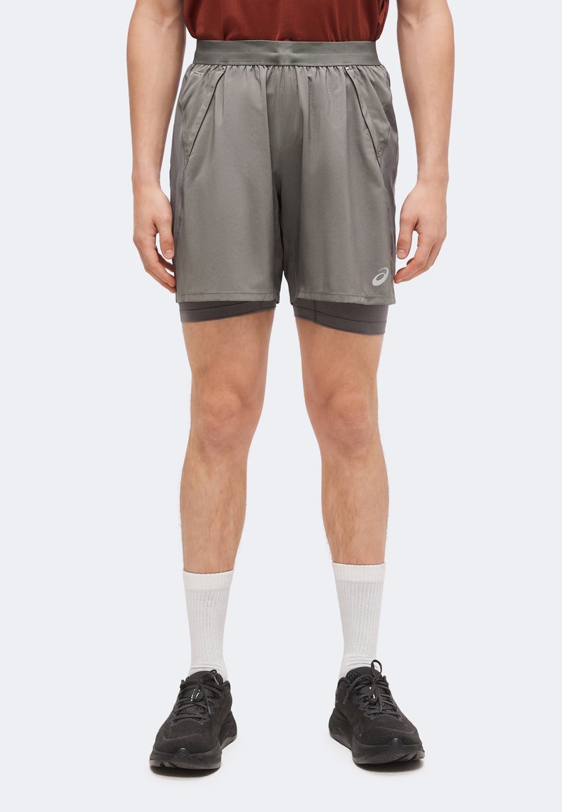 Homme portant un short de sport gris superposé, des chaussettes blanches montantes et des chaussures de course noires, debout droit devant un fond uni.