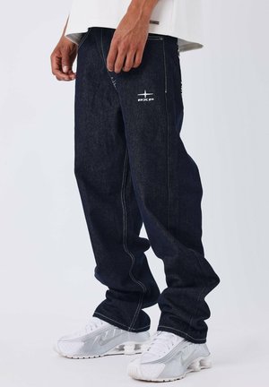 Persona que lleva jeans anchos de color azul oscuro con costuras blancas y un logo blanco "PXP", combinados con zapatillas Nike blancas con detalles plateados.