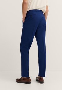 Blaue maßgeschneiderte Hose aus glattem Stoff, mit schlanker Passform, Gesäßtaschen und Knopfverschluss. Kombiniert mit braunen Lederschuhen.