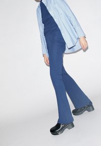 Pantalones acampanados de color navy con una textura acanalada, combinados con una camisa a rayas azul claro y zuecos negros con tachuelas plateadas en la suela.