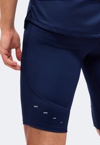 Gros plan d’une personne portant un short de sport ajusté bleu marine avec des bandes réfléchissantes sur la cuisse extérieure et un t-shirt bleu marine assorti.