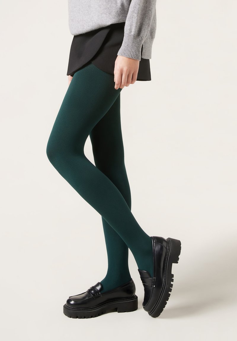 Femme portant des collants opaques vert foncé, des mocassins noirs, une jupe noire et un pull gris, debout devant un fond clair.