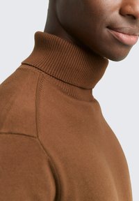Pull à col roulé en tricot marron avec un col côtelé et des coutures nettes, présenté sur un fond gris clair. Texture douce et design ajusté.