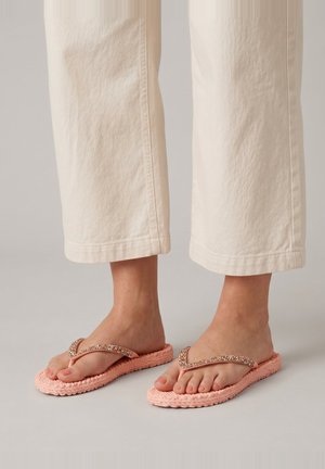 Pies con chanclas trenzadas de color rosa claro con tiras decorativas, combinadas con pantalones cortos de color crema sobre un fondo neutro.