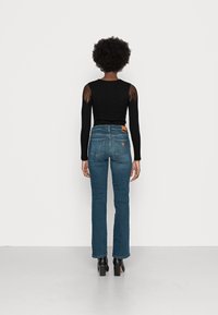 Top negro de manga larga con textura acanalada y paneles transparentes en los hombros, combinado con unos jeans de mezclilla azul ajustados y de pierna acampanada y botas negras de tacón. Etiqueta de cuero marrón en los jeans.