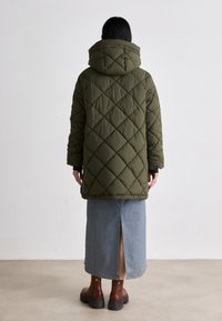 Olijfgroene gewatteerde pufferjas met een capuchon, voorzien van een ruitpatroon, gedragen over een spijkerrok met een achteropening en bruine enkellaarzen.