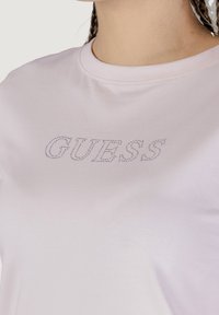 Lichtroze katoenen t-shirt met een ronde hals en korte mouwen; heeft "GUESS" in kleine strasssteentjes op de voorkant.