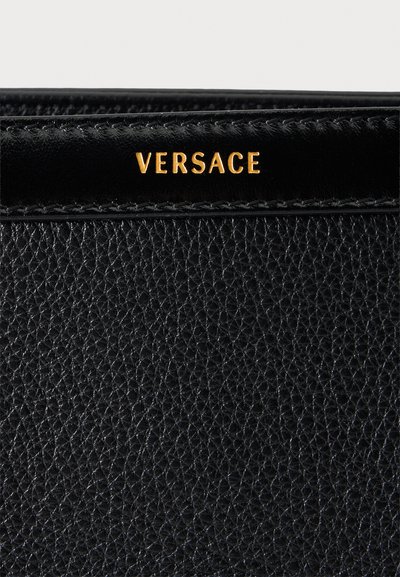 Surface en cuir noir texturé avec bord cousu et lettrage doré épelant "VERSACE" sur une bande près du haut.