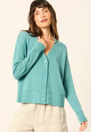 Frau trägt eine türkisfarbene Strickjacke mit Knöpfen und beige Hose, berührt eine goldene Halskette, vor einem schlichten hellen Hintergrund.