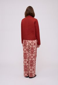 Giacca trapuntata rossa abbinata a pantaloni crema con un motivo floreale rosso ripetuto. L'outfit ha una vestibilità comoda e una consistenza morbida.