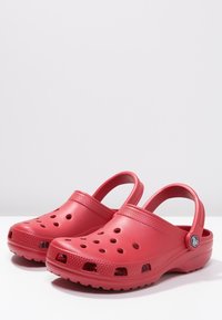 Paire de sabots rouges à enfiler avec des trous d’aération, des semelles texturées et des brides de talon réglables exposée sur une surface blanche.