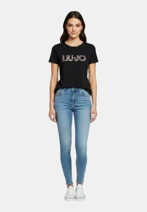 Donna in piedi che indossa una maglietta nera Liu Jo con logo floreale, jeans skinny azzurri e sneakers bianche su sfondo bianco.