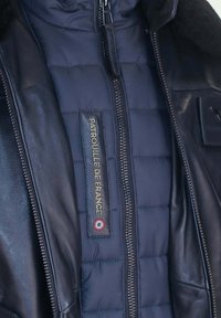 Doudoune bleu marine avec un extérieur en cuir, fermeture éclair à l'avant et un label brodé en or "Patrouille de France" avec un détail de rondel.
