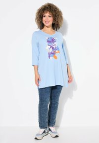 Tunika leggera blu con scollo tondo e maniche a tre quarti. Presenta una stampa grafica floreale in viola, arancione e blu. Abbinata a jeans scuri.