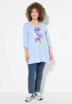 Helles blaues Tunika-Oberteil mit Rundhalsausschnitt und drei Viertelärmeln. Mit einem floralen Grafikdruck in Lila, Orange und Blau. Kombiniert mit dunklen Jeans.