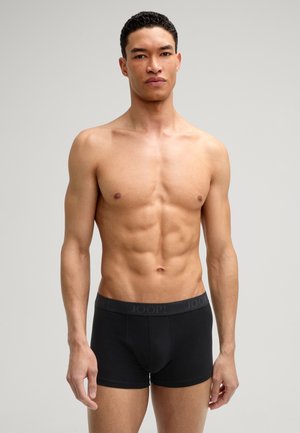 Hemdloser junger Mann mit kurzen lockigen Haaren, der schwarze Boxershorts trägt, steht vor einem einfarbigen hellgrauen Hintergrund.