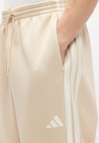 Beige joggers with a soft, smooth texture, featuring side pockets and three white stripes along the sides, accented with a white logo.