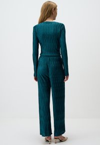 Jumpsuit lungo a maniche con texture a coste verticali in tonalità teal, pantaloni a gamba larga, vita aderente e tessuto morbido. Tacchi dorati completano il look.