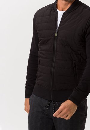 Homme portant une veste matelassée noire zippée sur une chemise blanche avec un pantalon à carreaux gris foncé, une main légèrement levée.