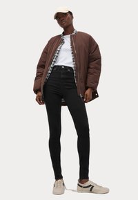 Chaqueta bomber marrón sobre una camisa blanca y una camisa de franela a cuadros, combinada con jeans skinny negros y zapatillas beige con acentos oscuros.