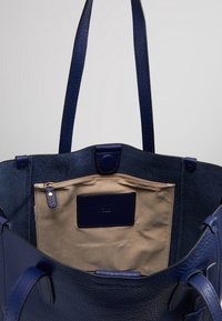 Suuri laivastonsininen nahkainen olkalaukku, jossa karkeahko pinta, ja jossa on beigen värinen sisäinen vetoketjutasku sekä kaksinkertainen kantokahvasuunnittelu.