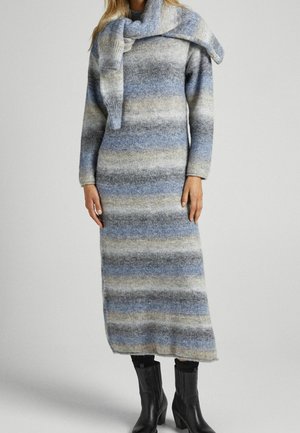 Strickkleid mit langen Ärmeln, das blau, grau und cremefarbene horizontale Streifen aufweist. Weicher Stoff, knielange Länge, getragen mit kniehohen schwarzen Stiefeln.