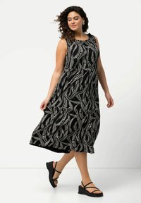 Schwarzes ärmelloses Kleid mit einem weißen Farnmuster, fließendem Design und Rüschenausschnitt. Kombiniert mit schwarzen Riemchensandalen mit Keilabsatz.