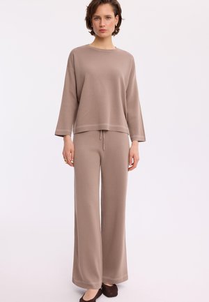 Kobieta w luźnej, długiej, taupe-bluzce i szerokich spodniach stoi na tle jednolitego jasnego tła.