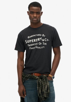 Camiseta de algodón negra con texto blanco de estilo vintage, combinada con jeans azules y una camisa de cuadros verde atada a la cintura. Detalle de reloj casual visible.
