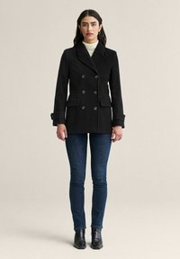 Svart dubbelknäppt kappa med stora knappar, sidofickor och uppklippt krage, kombinerad med mörka jeans och ankelstövlar.