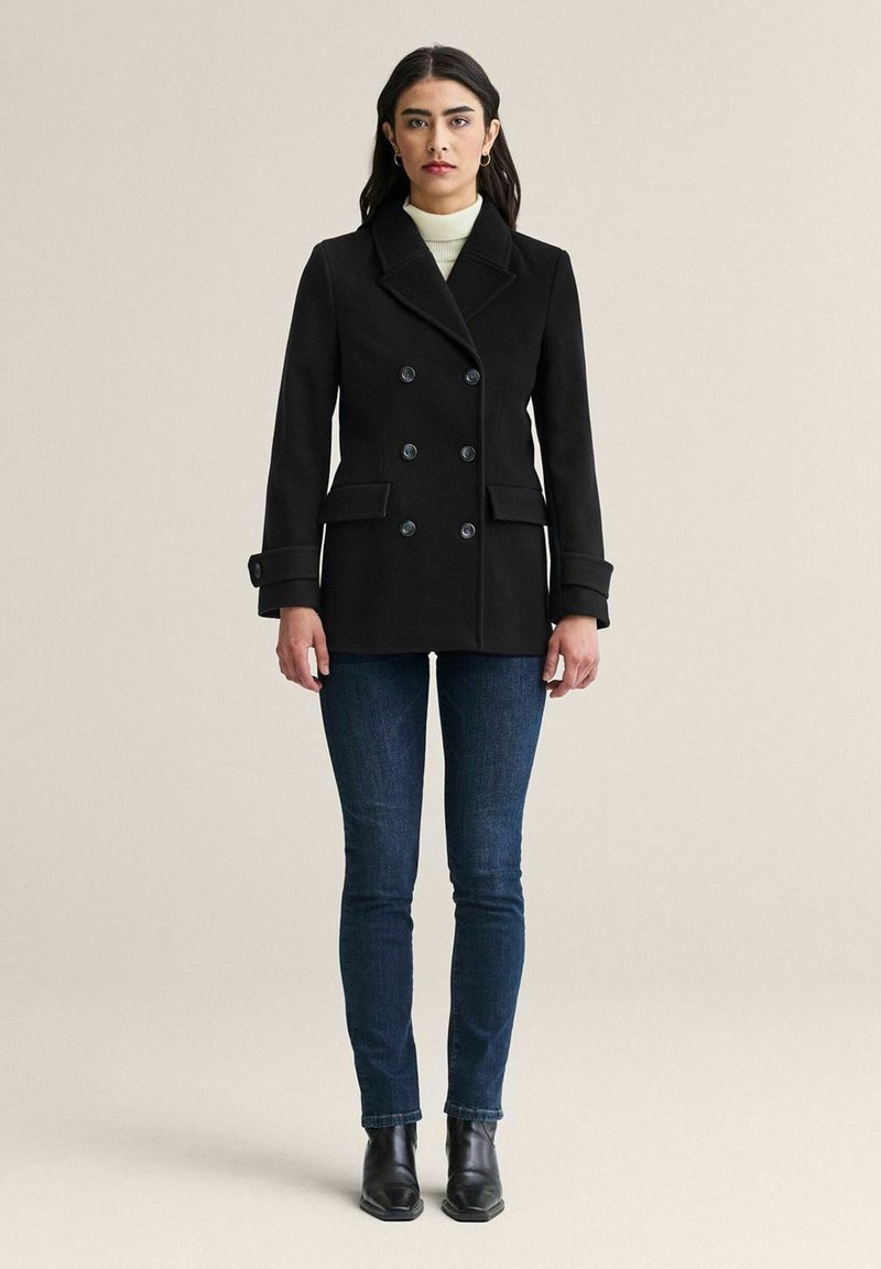 Svart dubbelknäppt kappa med stora knappar, sidofickor och uppklippt krage, kombinerad med mörka jeans och ankelstövlar.