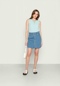 Blusa sem mangas azul clara com detalhe em laço, combinada com uma saia mini de ganga com botões frontais, e acessorizada com uma pequena bolsa branca e sandálias.