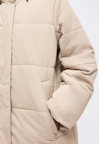 Veste d'hiver matelassée beige avec logo visible de la marque sur la poitrine et main glissée dans la poche latérale droite.