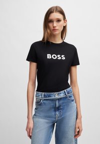 T-shirt noir en coton avec le grand texte blanc "BOSS" sur le devant, associé à un jean taille haute bleu clair. Design et coupe simples.