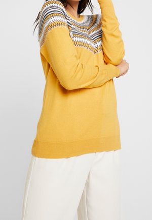 Femme portant un pull tricoté jaune à motifs géométriques blancs et noirs sur la poitrine, associé à un pantalon crème à jambes larges.