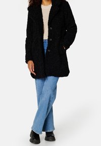 Schwarzer, flauschiger Mantel mit Reverskragen, Frontknöpfen und zwei seitlichen Taschen, kombiniert mit hellblauen, ausgestellt geschnittenen Jeans und schwarzen Stiefeln.