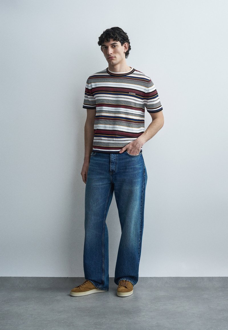 Young man standing against a plain wall wearing a striped short-sleeve shirt, blue wide-leg jeans, and brown sneakers.