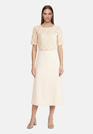 Femme portant un top court en dentelle crème à manches courtes et une jupe midi taille haute assortie, avec des sandales beige à talons carrés sur fond blanc.