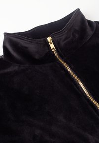 Zwarte fluwelen jas met een hoge kraag en een zichtbare goudkleurige rits die gedeeltelijk dicht is op een witte achtergrond.