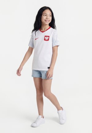 Djevojka dugom crnom kosom u bijelom dresu poljske nogometne reprezentacije, svijetlim traper kratkim hlačama i bijelim tenisicama, hoda gledajući udesno s blagim osmijehom.