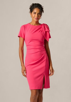 Femme aux cheveux bouclés portant une robe rose vif ajustée, à manches courtes, avec un détail nœud à l'épaule et une fente devant, debout sur un fond uni.