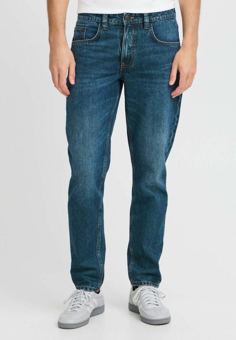 Dunkelblaue Jeans mit geradem Bein, fünf Taschen und subtiler Verwaschung, kombiniert mit weißen Sneakers vor einem schlichten Hintergrund.