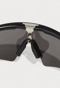 Schwarze Sonnenbrille mit schmalem Rahmen, grauen Gläsern, Gummibügeln und metallischen Akzenten an der Brücke. Glatte Oberfläche.