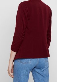Giacca aderente bordeaux con maniche lunghe, texture liscia, dettagli di arricciatura sui gomiti, abbinata a jeans azzurri chiari.