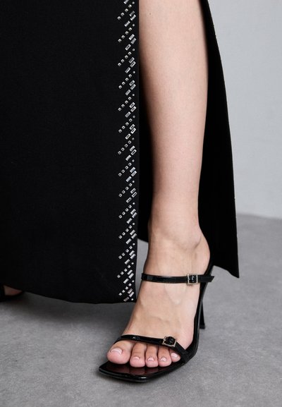 Rochie neagră cu embellismente geometrice argintii și o deschidere pe lateral. Sandale negre cu toc înalt, cu curea subțire și vârf pătrat.