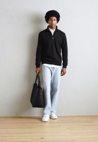 Black zippered sweater, white shirt beneath, light blue jeans, white sneakers, holding a black tote bag, standing on wooden floor against a gray wall.