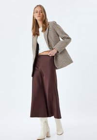 Blazer a scacchi marrone e crema, top bianco, gonna fluida in satin marrone e stivali bianchi con tacco a blocco al ginocchio. Motivi e strati testurizzati.