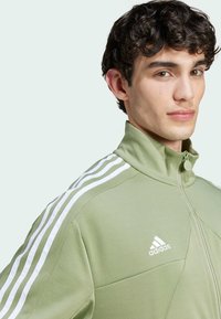 Giacca zip-up verde chiaro con colletto alto, caratterizzata da tre strisce bianche sulle spalle e un logo bianco sul petto. Tessuto morbido.