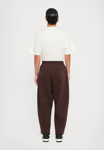 Brown, loose-fit sweatpants with elastic waistband and back pocket; worn with a cream short-sleeve shirt and black sneakers.